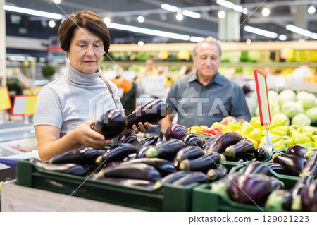 Elderly buyer woman chooses ripe eggplant on grocery store window Elderly buyer woman chooses ripe eggplant on grocery store window 129021223