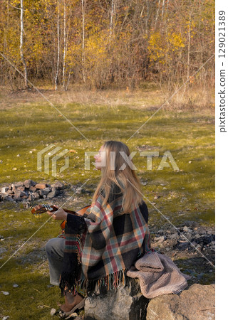 Blonde woman playing string guitar outdoors in autumn forest. Concept of sound therapy, mental health and wellness rituals. Calmness tranquility audio-sensory practices. Aura farming energy 129021389