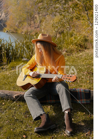 Portrait of smiling caucasian artist plays wooden acoustic guitar in autumnal park. Young songwriter plays string musical instrument hobby outside in nature fall time. Audio music healing Portrait of smiling caucasian artist plays wooden acoustic guitar in autumnal park. Young songwriter plays string musical instrument hobby outside in nature fall time. Audio music healing 129021400