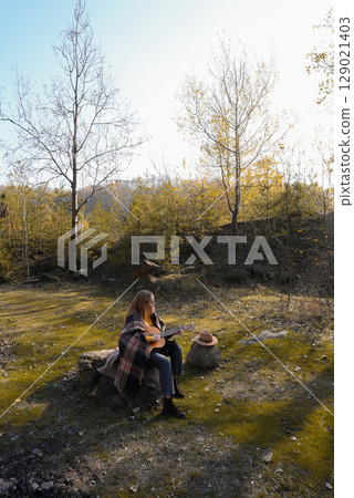 Beautiful blonde girl playing acoustic guitar and singing relaxing enjoying outdoors on sunny autumn day. Hippie woman in boho style learning and practicing playing musical instrument. Fall leaves 129021403