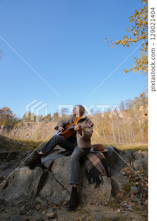 Beautiful blonde girl playing acoustic guitar and singing relaxing enjoying outdoors on sunny autumn day. Hippie woman in boho style learning and practicing playing musical instrument. Fall leaves Beautiful blonde girl playing acoustic guitar and singing relaxing enjoying outdoors on sunny autumn day. Hippie woman in boho style learning and practicing playing musical instrument. Fall leaves 129021404