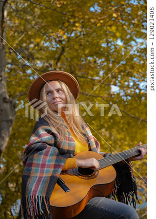 Beautiful blonde girl playing acoustic guitar and singing relaxing enjoying outdoors on sunny autumn day. Hippie woman in boho style learning and practicing playing musical instrument. Fall leaves 129021408