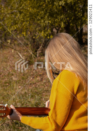 Beautiful blonde girl playing acoustic guitar and singing relaxing enjoying outdoors on sunny autumn day. Hippie woman in boho style learning and practicing playing musical instrument. Fall leaves Beautiful blonde girl playing acoustic guitar and singing relaxing enjoying outdoors on sunny autumn day. Hippie woman in boho style learning and practicing playing musical instrument. Fall leaves 129021411