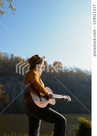 Blonde woman playing string guitar outdoors in autumn forest. Concept of sound therapy, mental health and wellness rituals. Calmness tranquility audio-sensory practices. Aura farming energy 129021417
