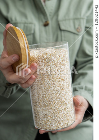 Unrecognizable woman holding refillable glass jar with raw fine ground barley porridge. Refill product format eco-friendly living. Filling reusable container Whole spelt grain species. High protein 129021442