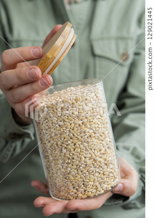 Unrecognizable woman holding refillable glass jar with raw pearl barley porridge. Refill product format eco-friendly living. Filling reusable container Whole spelt grain species. High protein fiber Unrecognizable woman holding refillable glass jar with raw pearl barley porridge. Refill product format eco-friendly living. Filling reusable container Whole spelt grain species. High protein fiber 129021445