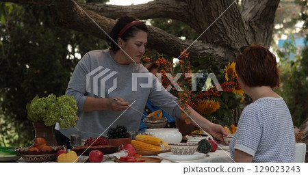 Guests enjoy Thanksgiving dinner outdoors with an array of colorful dishes displayed on a rustic table. Lush vegetables and fruits create a festive atmosphere filled with warmth and gratitude. 129023243