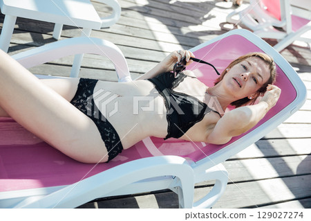 Woman in black swimsuit sunbathing on sun lounger on beach Woman in black swimsuit sunbathing on sun lounger on beach 129027274