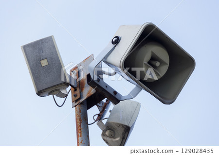 A Multifaceted Array of Outdoor Speakers Mounted on a Metal Pole against a Clear Blue Sky, Showcasing the Design and Functionality of Public Address Systems in Urban Areas 129028435