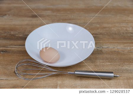 A Simple Culinary Setup: An Egg and a Whisk on a Plate Ready for Cooking or Baking in a Clean Kitchen Environment 129028537
