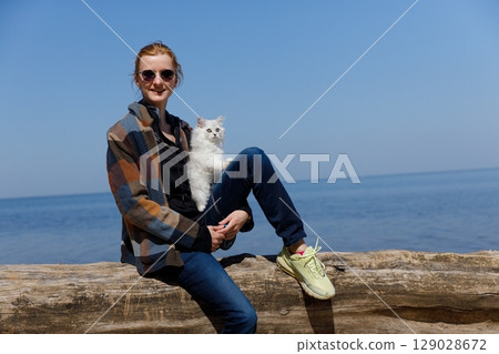 A woman sitting by the water with her fluffy cat on a sunny day, enjoying nature's beauty and capturing a moment of friendship and joy by the serene lakeside. A woman sitting by the water with her fluffy cat on a sunny day, enjoying nature's beauty and capturing a moment of friendship and joy by the serene lakeside. 129028672