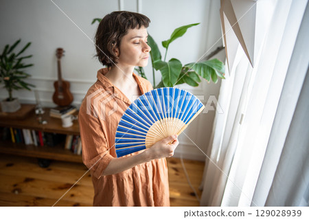 Exhausted sweaty woman fanning herself looking out window suffering from summer heat without AC Exhausted sweaty woman fanning herself looking out window suffering from summer heat without AC 129028939