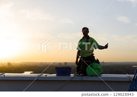 Safety and Accomplishment: Rooftop Worker Celebrates at Sunset with A Thumbs Up in Vibrant Yellow-Green Safety Gear Against the Background of a Dusk Cityscape Safety and Accomplishment: Rooftop Worker Celebrates at Sunset with A Thumbs Up in Vibrant Yellow-Green Safety Gear Against the Background of a Dusk Cityscape 129028943