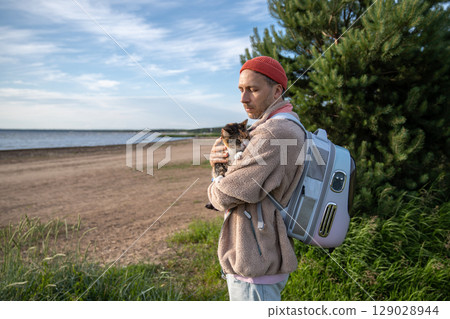 Loving man cuddling petting cat, look at sea coastline, backpack carrier for animal travel comfort 129028944