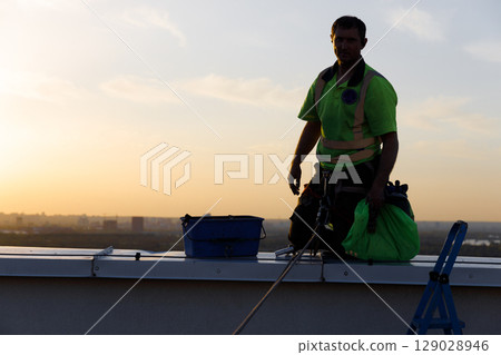 A Rooftop Worker in a Safety Harness at Dusk, Showcasing the Courageous and Daring Work of High-Rise Maintenance and Cleaning in Urban Landscapes A Rooftop Worker in a Safety Harness at Dusk, Showcasing the Courageous and Daring Work of High-Rise Maintenance and Cleaning in Urban Landscapes 129028946