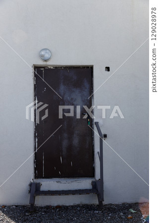 An Old, Weathered Brown Door with a Small Light Fixture Above and an Adjacent Step on a Simple White Wall Surrounded by Small Pebbles Reflecting a Rustic Atmosphere and Character 129028978
