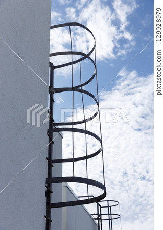 Modern Architectural Elements with an Intriguing Spiral Fire Escape Ladder Against a Blue Sky with Fluffy White Clouds Capturing Minimalist Design Aesthetics 129028979