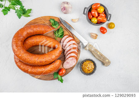 Sliced smoked sausage on a wooden board with fresh herbs, mustard, garlic, tomatoes, and a rustic knife on a light background. top view. copy space 129029093