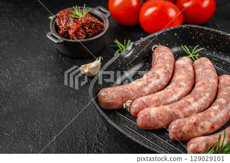 Raw pork sausages seasoned with herbs and spices arranged in a grill pan with garlic, tomatoes, and sun-dried tomatoes on a black textured background. copy space, top view. Raw pork sausages seasoned with herbs and spices arranged in a grill pan with garlic, tomatoes, and sun-dried tomatoes on a black textured background. copy space, top view. 129029101