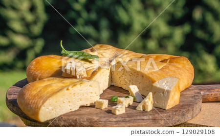 A round wheel of smoked cheese with cut wedges and cubes is displayed on a rustic wooden board outdoors, surrounded by greenery and sunlight 129029108