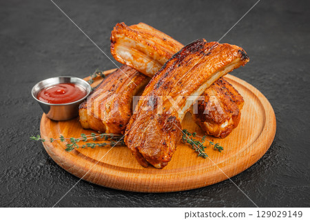 Grilled pork ribs stacked on a wooden board with fresh herbs and a metal cup of ketchup on a dark textured background Grilled pork ribs stacked on a wooden board with fresh herbs and a metal cup of ketchup on a dark textured background 129029149