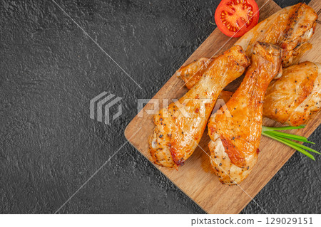 Juicy roasted chicken drumsticks served on a wooden board with tomato and green onion garnish on a dark background. top view. copy space Juicy roasted chicken drumsticks served on a wooden board with tomato and green onion garnish on a dark background. top view. copy space 129029151
