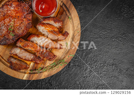 Sliced grilled pork steak served with barbecue sauce and herbs on a rustic wooden plate over a dark background. top view. copy space Sliced grilled pork steak served with barbecue sauce and herbs on a rustic wooden plate over a dark background. top view. copy space 129029156