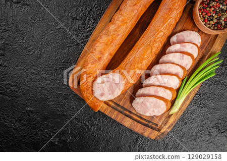 Smoked sausage sliced and served on a wooden board with green onions and a bowl of colorful peppercorns on a dark background. top view. copy space. 129029158
