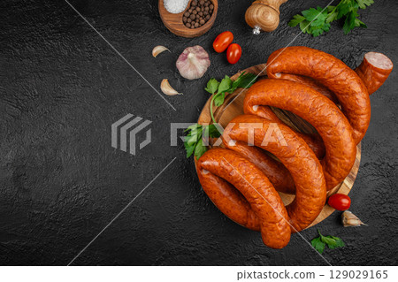 smoked sausages on a wooden board with herbs, garlic, pepper, and tomatoes on a dark textured background. top view. copy space smoked sausages on a wooden board with herbs, garlic, pepper, and tomatoes on a dark textured background. top view. copy space 129029165
