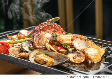 A hot iron skillet sizzles with a vibrant seafood medley including octopus, mussels, clams, shrimp, scallops, tomatoes, and asparagus in a rich savory sauce. A hot iron skillet sizzles with a vibrant seafood medley including octopus, mussels, clams, shrimp, scallops, tomatoes, and asparagus in a rich savory sauce. 129029168