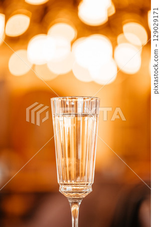 A tall crystal glass filled with sparkling white wine stands elegantly against a warm bokeh-lit background in a cozy, refined setting 129029171