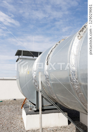 Large Industrial Ventilation System Featuring a Silver Duct with Insulation and a Control Unit, Set Against a Bright Blue Sky with Clouds 129029661