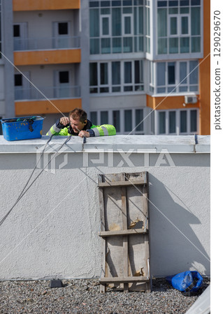 A Rooftop Worker: Skilled Technician Performing Maintenance on a Building Roof with Modern Residential Units in the Background Under Clear Blue Sky 129029670