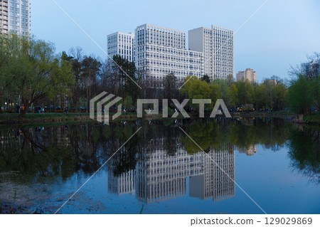 Stunning View of Modern Architecture Reflected in a Serene Pond Surrounded by Lush Greenery and People Enjoying the Evening Ambience in a Vibrant Urban Park Setting 129029869