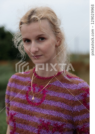 A Portrait of Elegance: A Young Woman with Beautiful Curly Hair Wearing a Stylish Striped Sweater in Earthy Tones Against a Softly Blurred Natural Background 129029968