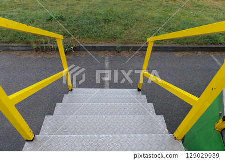 A Close-Up View of Yellow Railings and Metal Steps Leading Down from a Structure onto an Asphalt Path with Grass and Wildflowers in the Background, Creating a Curious Scene A Close-Up View of Yellow Railings and Metal Steps Leading Down from a Structure onto an Asphalt Path with Grass and Wildflowers in the Background, Creating a Curious Scene 129029989