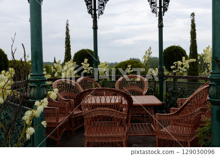 Charming Outdoor Seating Area with Wicker Furniture Surrounded by Lush Greenery and Beautiful White Flowers Under a Gray Sky Charming Outdoor Seating Area with Wicker Furniture Surrounded by Lush Greenery and Beautiful White Flowers Under a Gray Sky 129030096