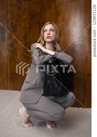 A stylish young woman, long blonde hair, poses in a grey suit, holding a black leather bag against a warm wooden background 129031126