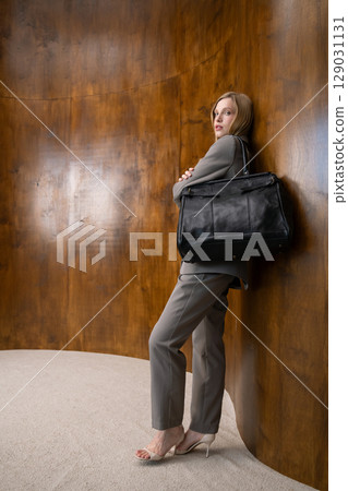 A stylish young woman, long blonde hair, poses in a grey suit, holding a black leather bag against a warm wooden background A stylish young woman, long blonde hair, poses in a grey suit, holding a black leather bag against a warm wooden background 129031131