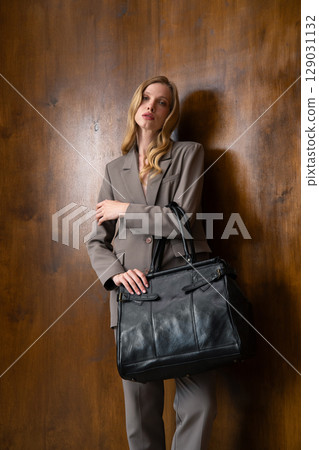 A stylish young woman, long blonde hair, poses in a grey suit, holding a black leather bag against a warm wooden background A stylish young woman, long blonde hair, poses in a grey suit, holding a black leather bag against a warm wooden background 129031132