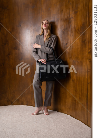 A stylish young woman, long blonde hair, poses in a grey suit, holding a black leather bag against a warm wooden background 129031133