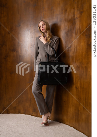 A stylish young woman, long blonde hair, poses in a grey suit, holding a black leather bag against a warm wooden background 129031142