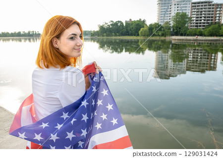 Young womanl holding USA national flag on her shoulders with high office buildings in a city celebrating United States independence day. 129031424