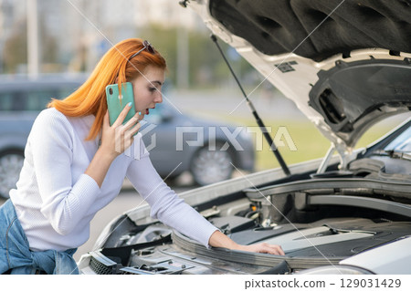 Young woman standing near broken car with popped hood talking on her mobile phone while waiting for help. Young woman standing near broken car with popped hood talking on her mobile phone while waiting for help. 129031429