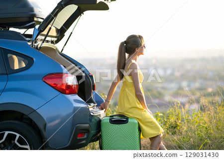 Young woman resting on a green suitcase near her car in summer nature. Travel and vacations concept. 129031430