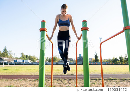 Young sportive athlete woman doing fitness exercise on metal bars outdoors at stadium court. Young sportive athlete woman doing fitness exercise on metal bars outdoors at stadium court. 129031452