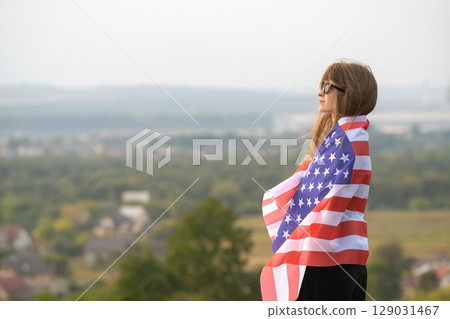 Young happy woman with long hair holding waving on wind american national flag on her sholders resting outdoors enjoying warm summer day. 129031467