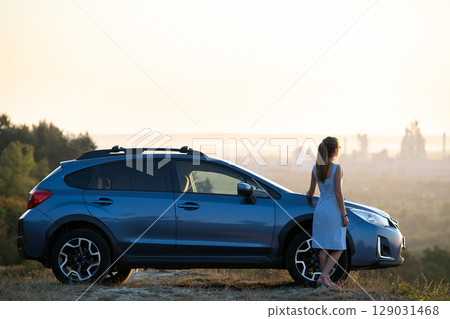 Young female driver resting near her car enjoying warm summer evening. Travel and getaway concept. 129031468
