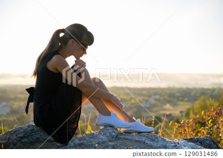 Young depressed woman in black short summer dress sitting on a rock thinking outdoors at sunset. Fashionable female contemplating in warm evening in nature. Young depressed woman in black short summer dress sitting on a rock thinking outdoors at sunset. Fashionable female contemplating in warm evening in nature. 129031486