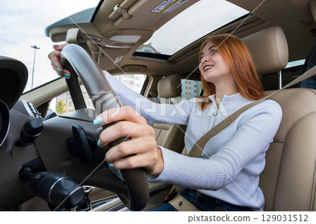 Wide angle view of young redhead woman driver fastened by seatbelt driving a car smiling happily. 129031512